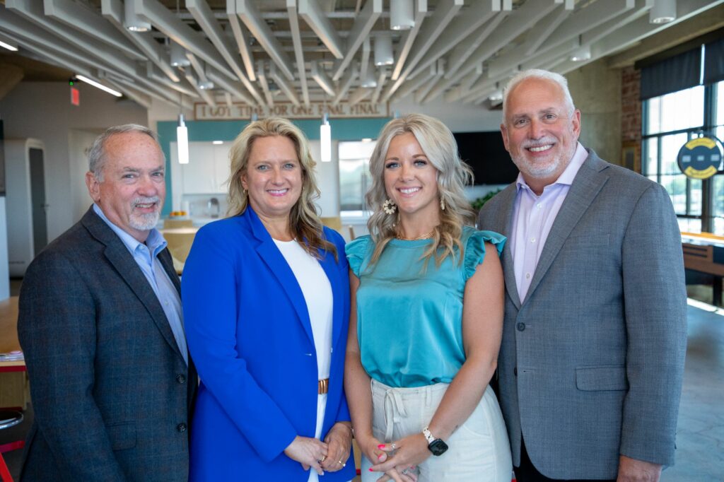 Four people are standing side by side in an office setting, smiling at the camera. The group includes two men and two women, all dressed in professional attire. The background features modern office decor with large windows and ample lighting.