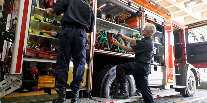 Two firefighters, both in uniforms, are organizing equipment in a fire truck. One is standing on the truck's step, reaching into a compartment filled with hoses and tools. The other is opening another compartment, also containing organized equipment.