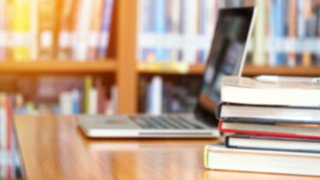 A wooden desks in a library with books and a laptop on top.