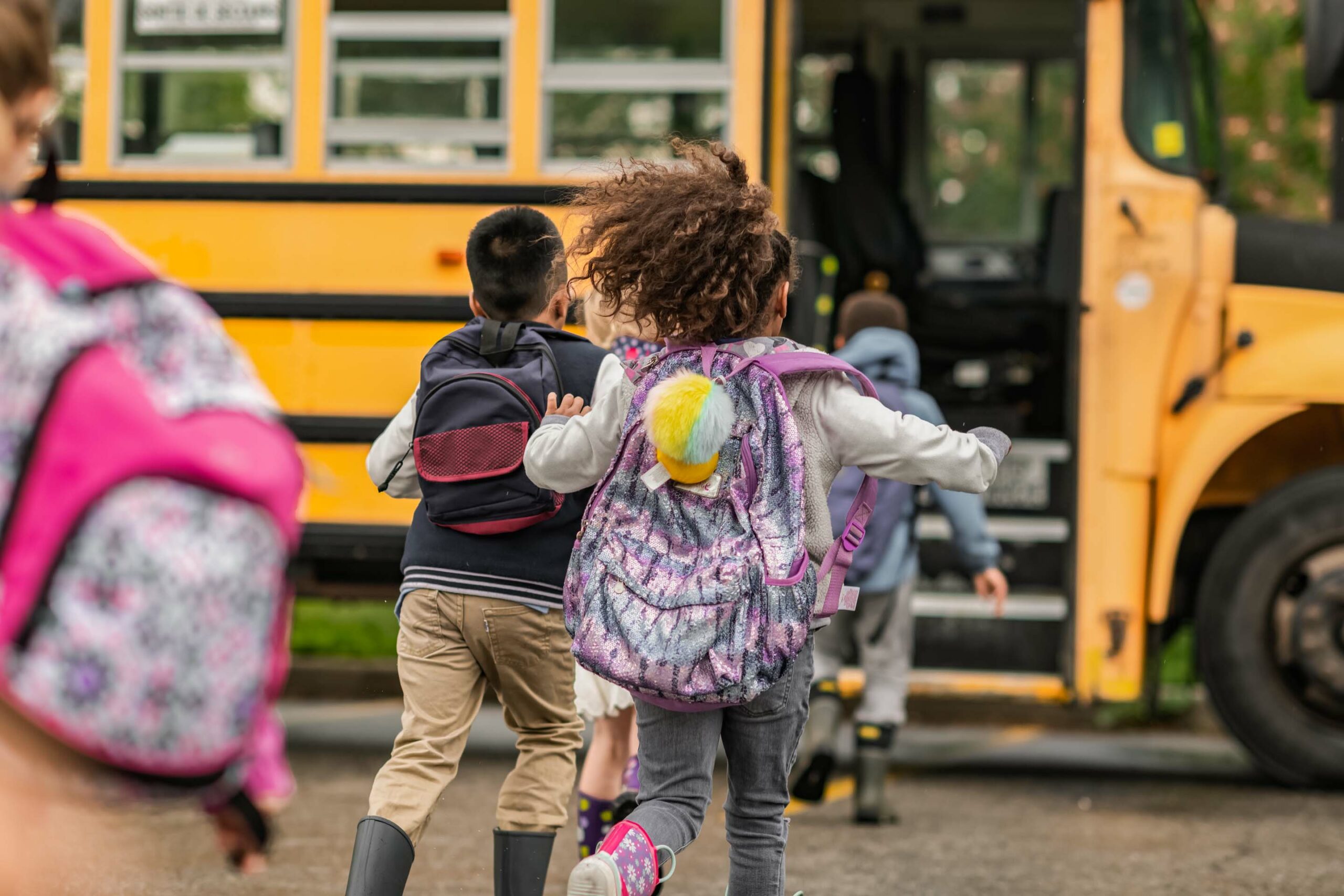 Children wearing backpacks are excitedly running towards a yellow school bus. The bus has an open door, and other children are seen boarding. The scene suggests a school day, with energetic movement from the group of kids.