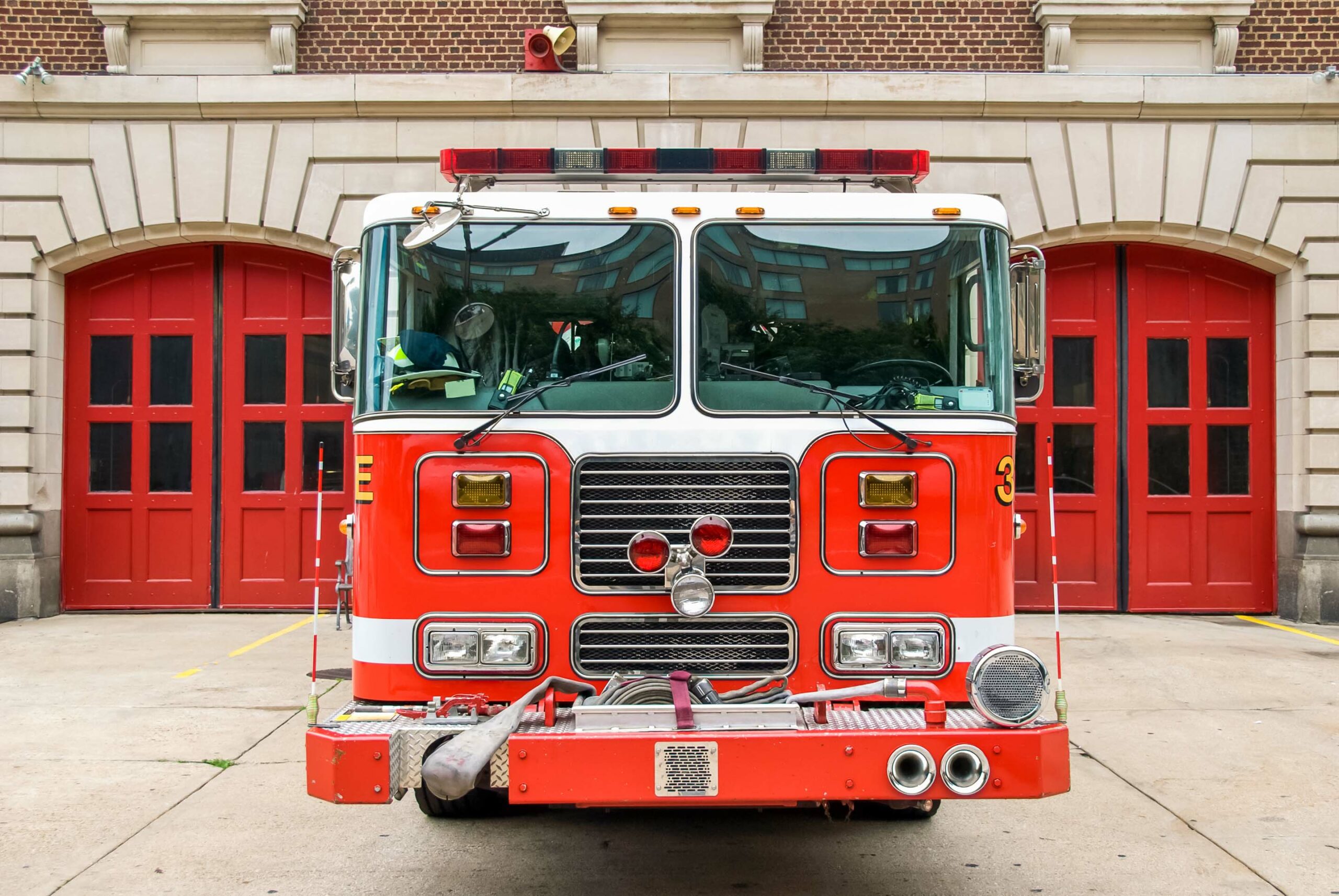 A red and white fire truck is parked directly in front of a fire station with red double doors. The fire truck features various equipment, lights, and hoses, with a symmetrical and bold front design. The building behind has beige stone and brick architecture.