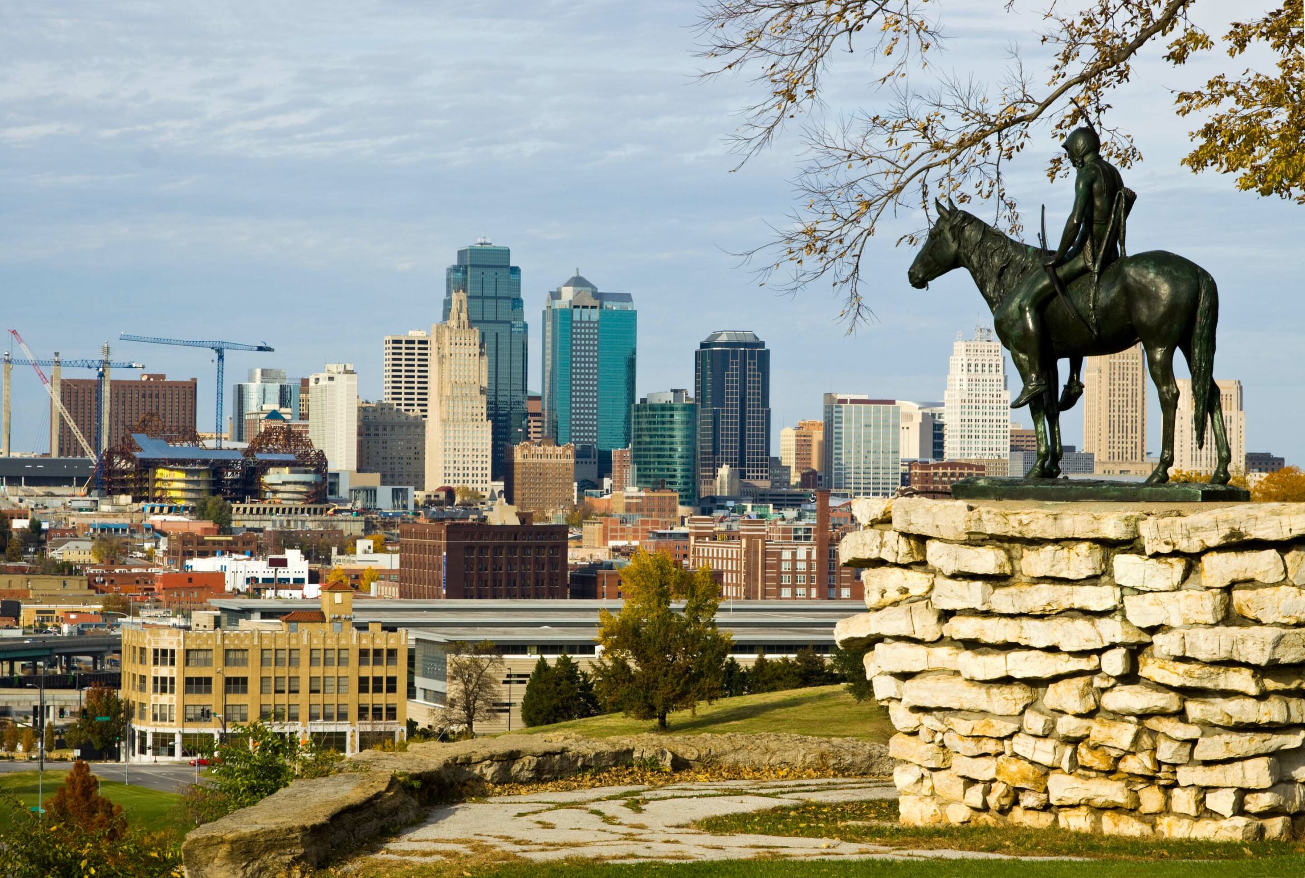 A bronze statue of a Native American on horseback overlooks the skyline of Kansas City. The cityscape features a mix of modern and historic buildings under a partly cloudy sky. The statue is perched on a stone pedestal surrounded by a grassy area.