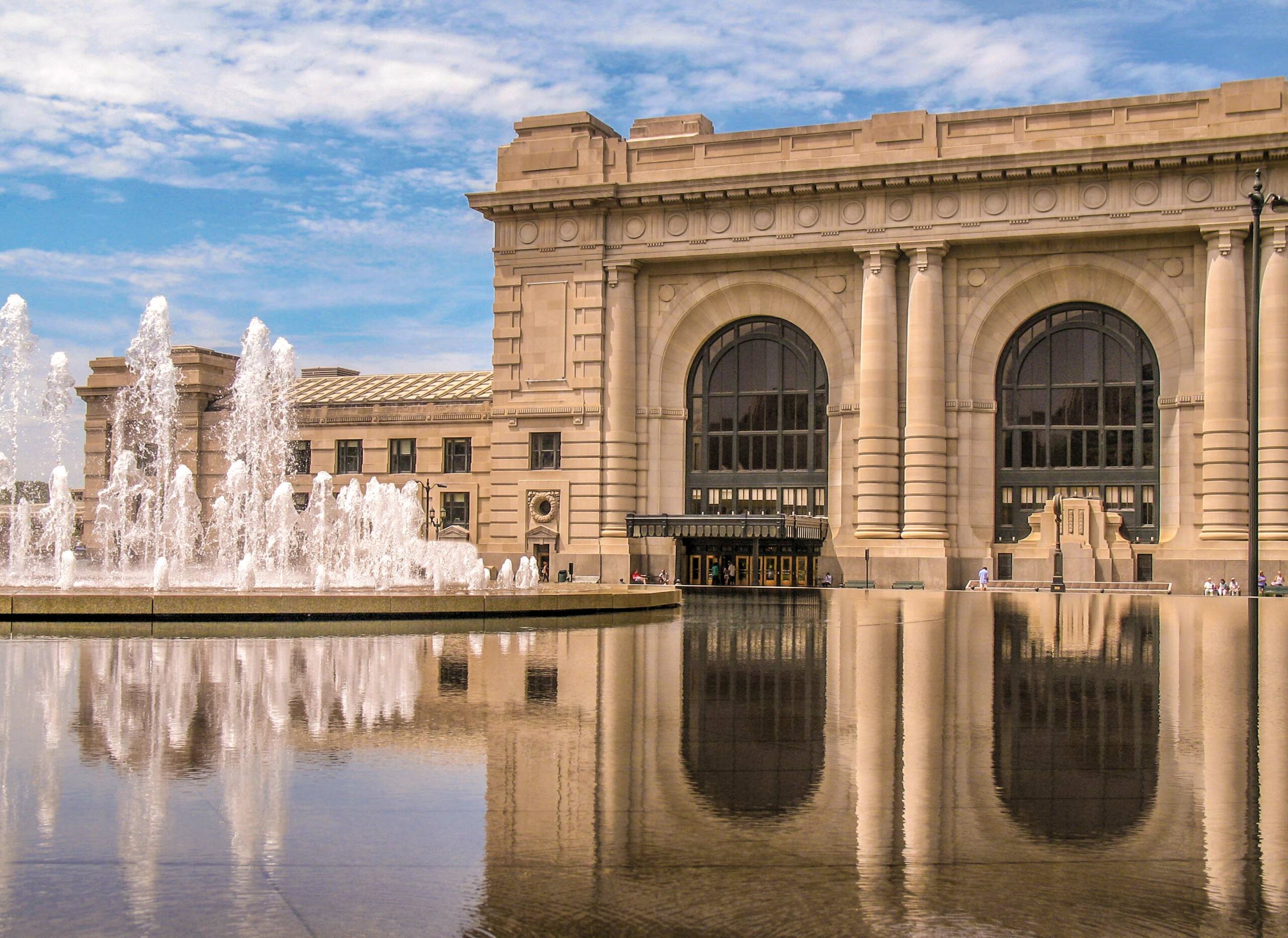 A grand, historic building with large arched windows is reflected in a calm pool of water. In front, a group of fountains sprays water into the air against a backdrop of a partly cloudy sky. The beige facade of the building contrasts with the blue sky and water.