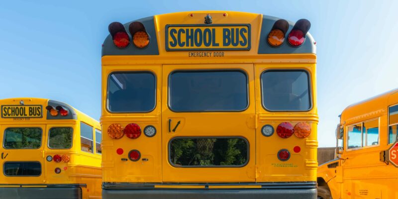 Rear view of a yellow school bus with distinct red and amber lights. The bus is parked next to a similar bus, and another is partially visible on the right. The scene is set under a clear blue sky.