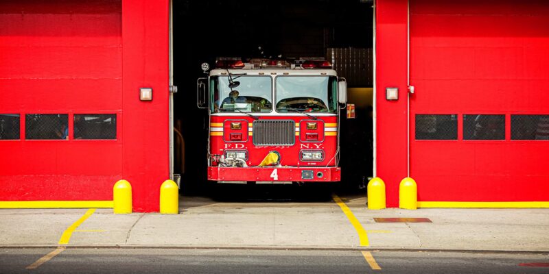 A red fire truck labeled FDNY is parked inside an open garage doorway of a fire station. The garage is painted red with two vertical rectangular windows on each side of the truck. The concrete sidewalk and street in front of the fire station are visible.