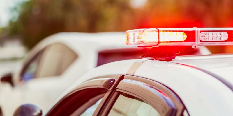 A close-up of a police car with its red emergency lights flashing. The car is parked beside another vehicle, with blurred trees in the background. The image suggests an active or recent traffic stop or emergency response.