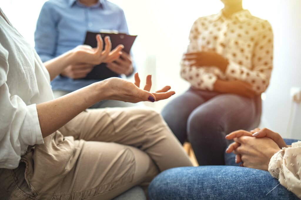 A group of people sitting in a circle during a discussion or therapy session. One person's hands are gesturing while another person holds a clipboard, possibly taking notes. The setting appears to be casual and focused on conversation or support.