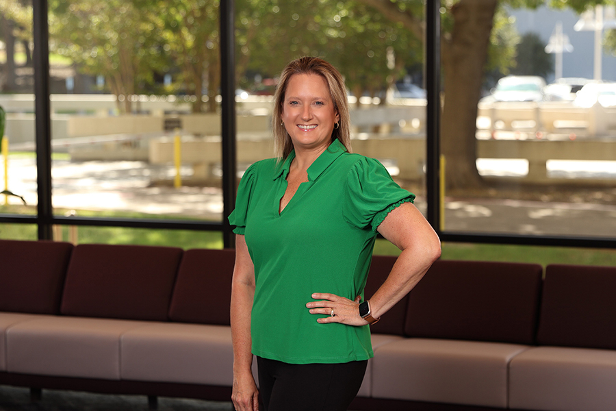 A person stands indoors wearing a green blouse and black pants, smiling with one hand on their hip. Large windows fill the background with a view of trees and a sunlit street.