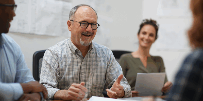 A middle-aged man wearing glasses smiles and gestures while talking during a meeting. Two colleagues sit nearby, one holding a tablet, and papers are spread out on the table. Drawings are pinned on the wall behind them.