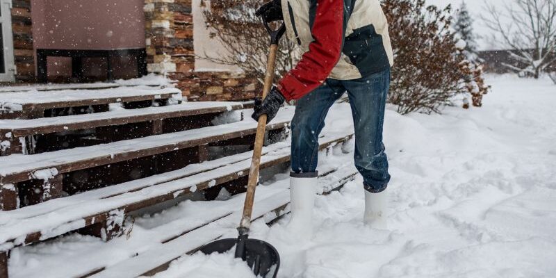 A person wearing winter clothing and white boots is shoveling snow in front of a house with steps, during a snowfall. Bushes and a snowy yard are visible in the background.