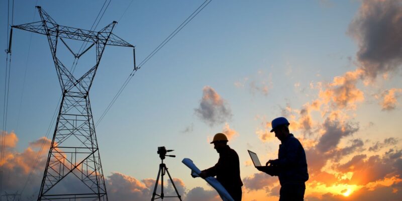 Two engineers in hard hats work near a tripod and power lines at sunset, one holding blueprints and the other using a laptop, with a dramatic sky and tall electricity pylons in the background.