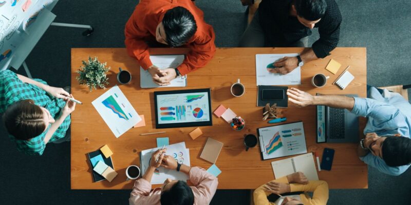 Six people sit around a wooden table with laptops, charts, coffee cups, and notes, having a collaborative meeting. Data graphs and documents are spread out as they discuss and share ideas from above.