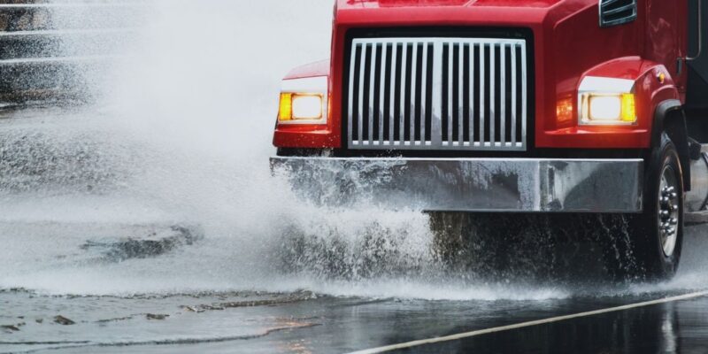 A red semi-truck drives through a large puddle on a wet road, creating a big splash of water as it moves forward.