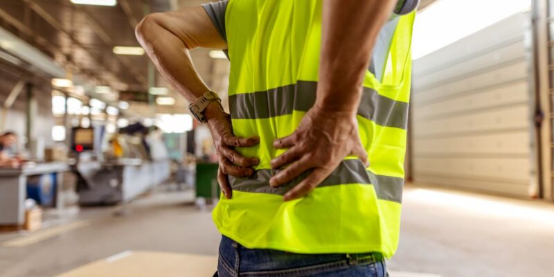 A worker in a high-visibility vest stands indoors holding their lower back in pain, suggesting discomfort or injury, with industrial equipment and materials visible in the background.