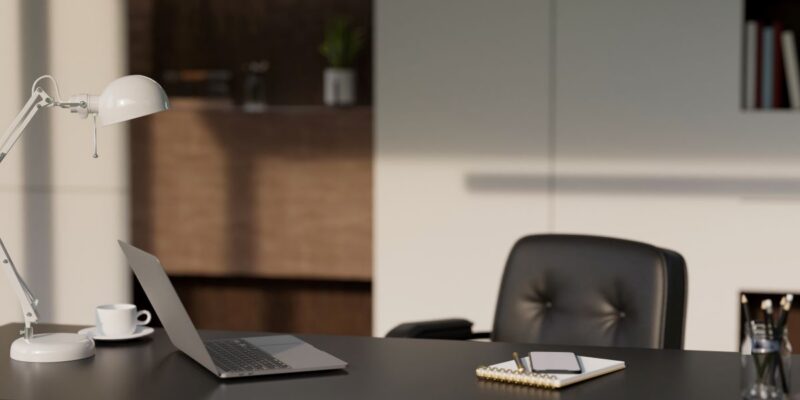 Modern office desk with a laptop, notepad, tablet, lamp, and coffee cup. A black chair is behind the desk. Shelves with books and plants are visible in the blurred background. Sunlight casts shadows in the room.